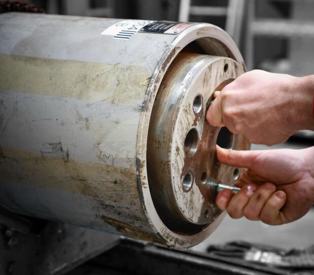 Eine Person schraubt mit einer Hand einen Bolzen in eine runde Metallplatte, die an einem großen Rohr befestigt ist